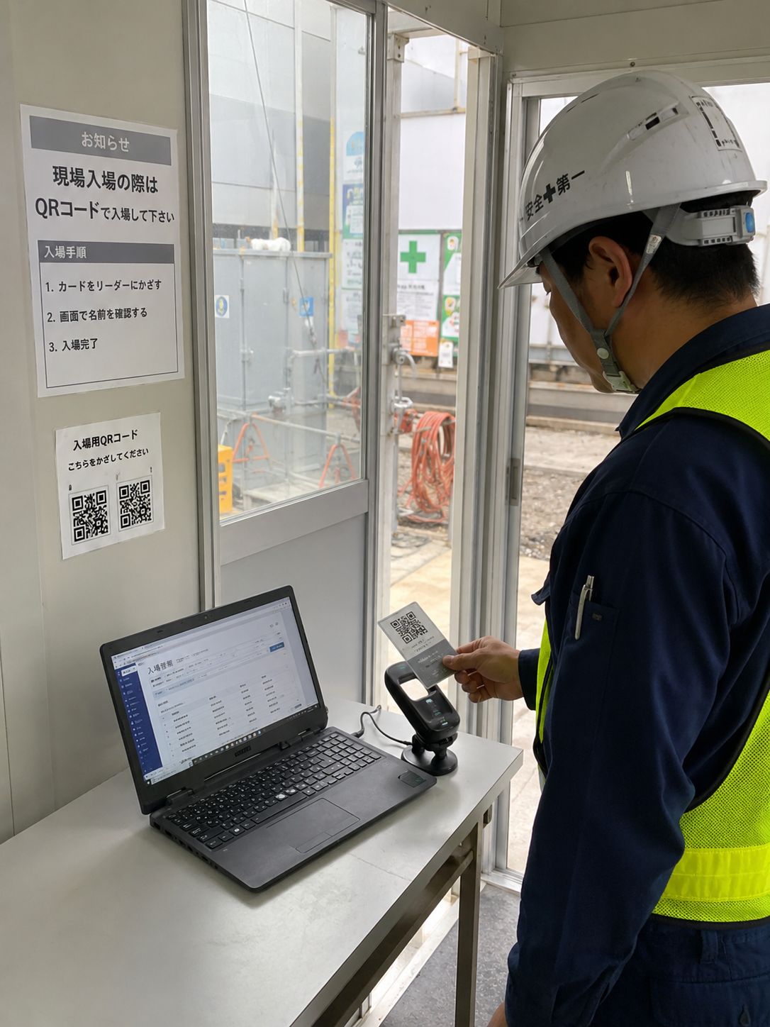 建設現場のプレハブ事務所入口で、ヘルメットをかぶった作業員がQRコード付きカードを市販バーコードリーダーにかざし、ノートPCのWeb管理画面に入場記録が登録されている写真品質のイメージ。大掛かりなゲートや顔認証専用機器は置かず、机上のPC・USBバーコードリーダー・QRカードだけで簡易設置している。画像内テキストは最小限。横長ワイド比率。